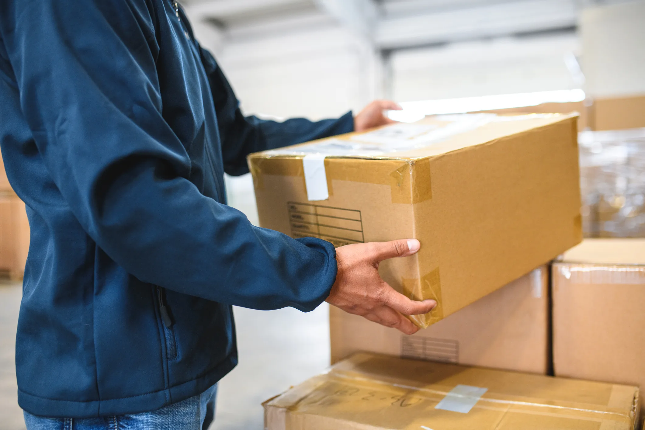 Asian delivery worker holding cardboard parcel, wearing blue uniform and standing inside of an airport warehouse.